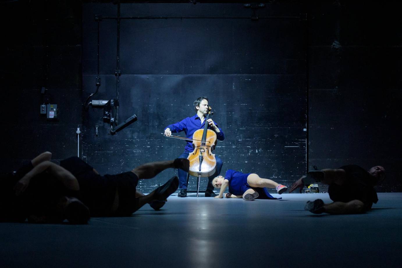 A cellist plays in light while the company lies on the floor